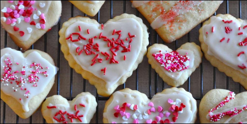 Heart-shaped cookies with white icing and red and pink sprinkles on a cooling rack.